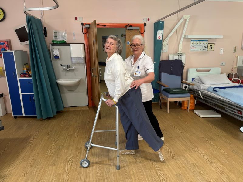 Jean Butcher standing with a frame and stretching alongside her physio, Ann.